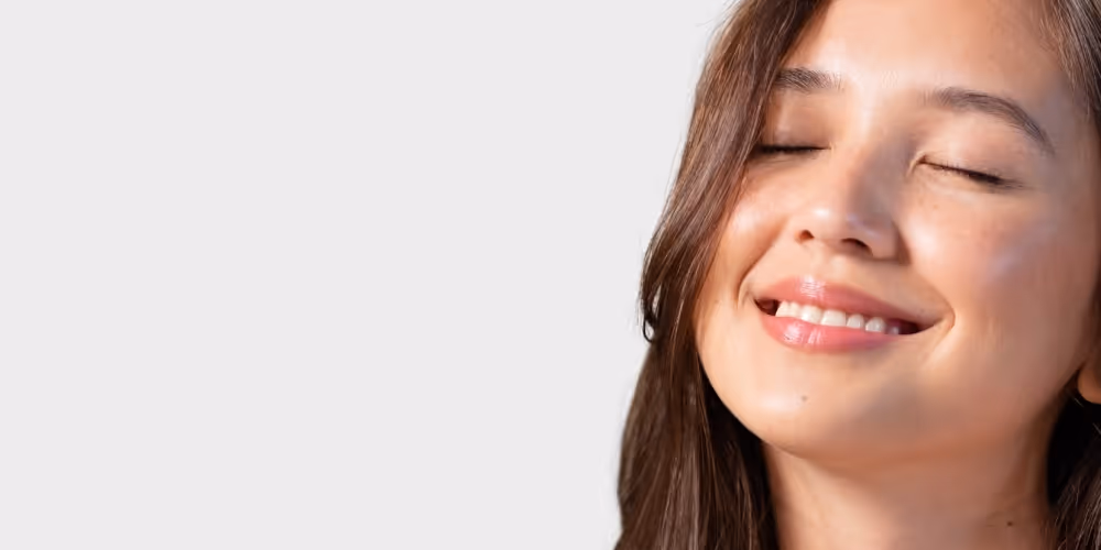 Close-up of a smiling woman with eyes closed and long brown hair against a plain gray background.