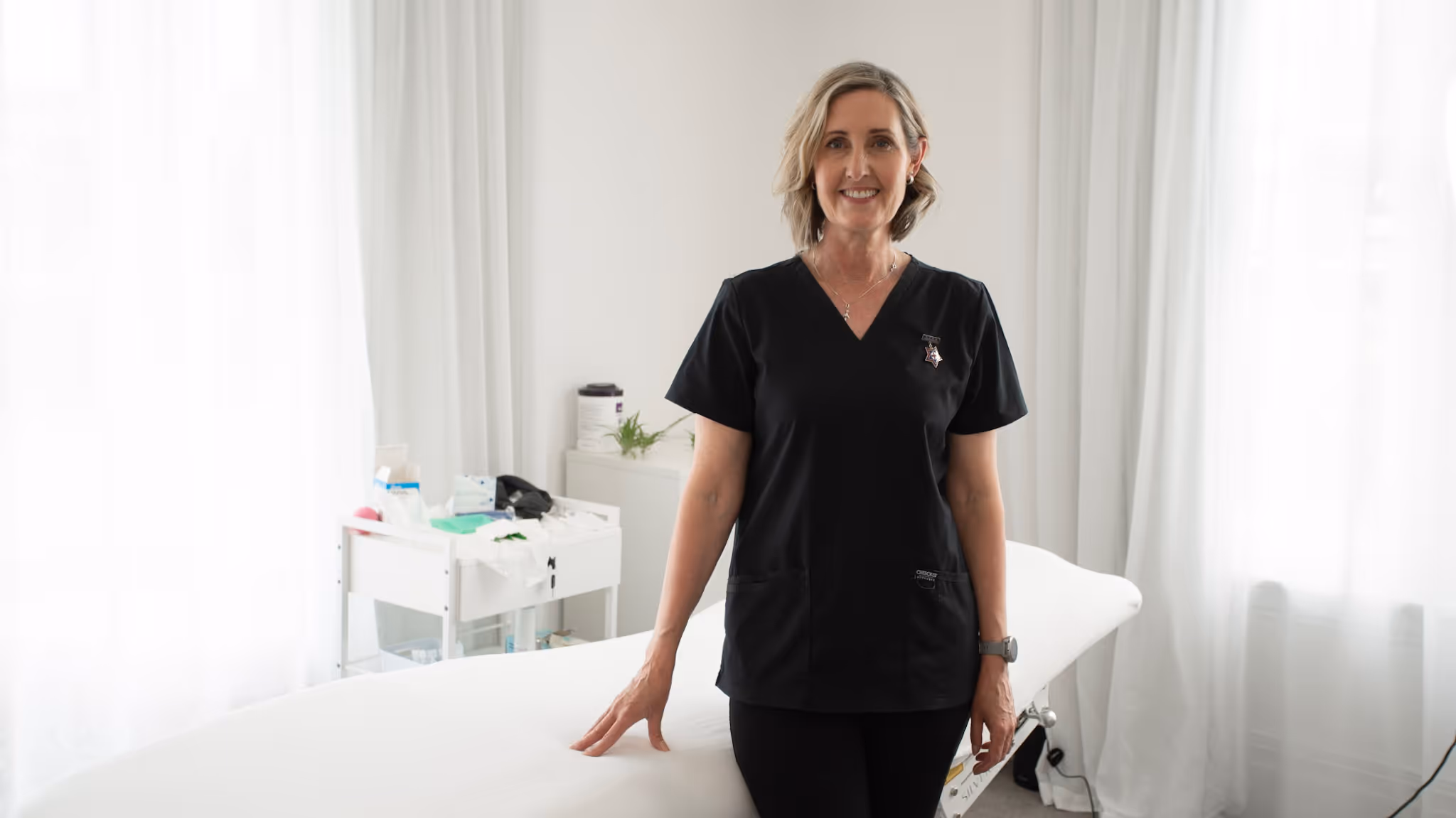 Andrea Harris smiling in black scrubs standing in her Aesthetic studio in The Vale, Merivale.