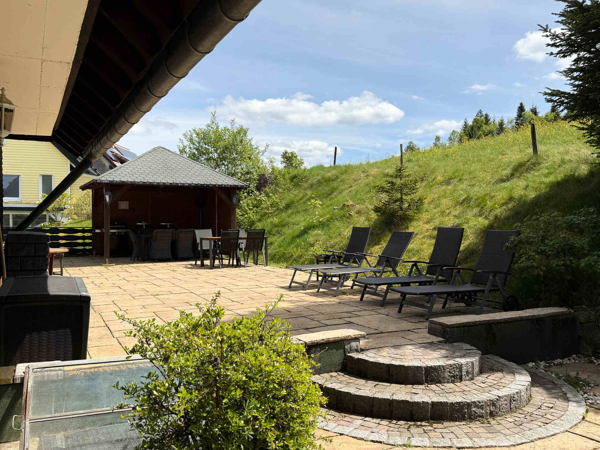 Sonnenterrasse mit Pavillon, Tischgruppe und mehreren Liegestuehlen an einem begruenten Hang