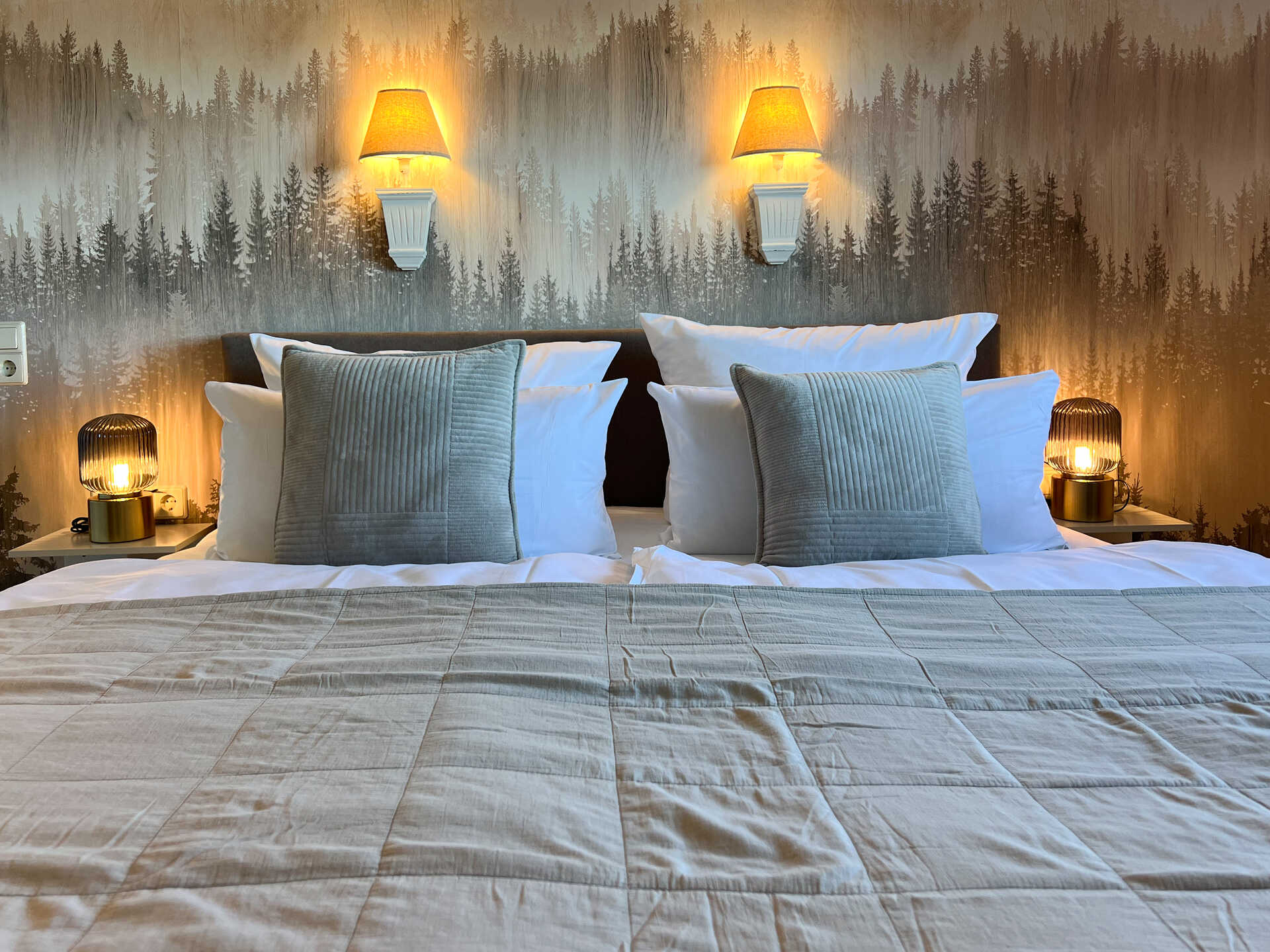 Bett mit Kissen und Nachttischlampen vor Waldtapete