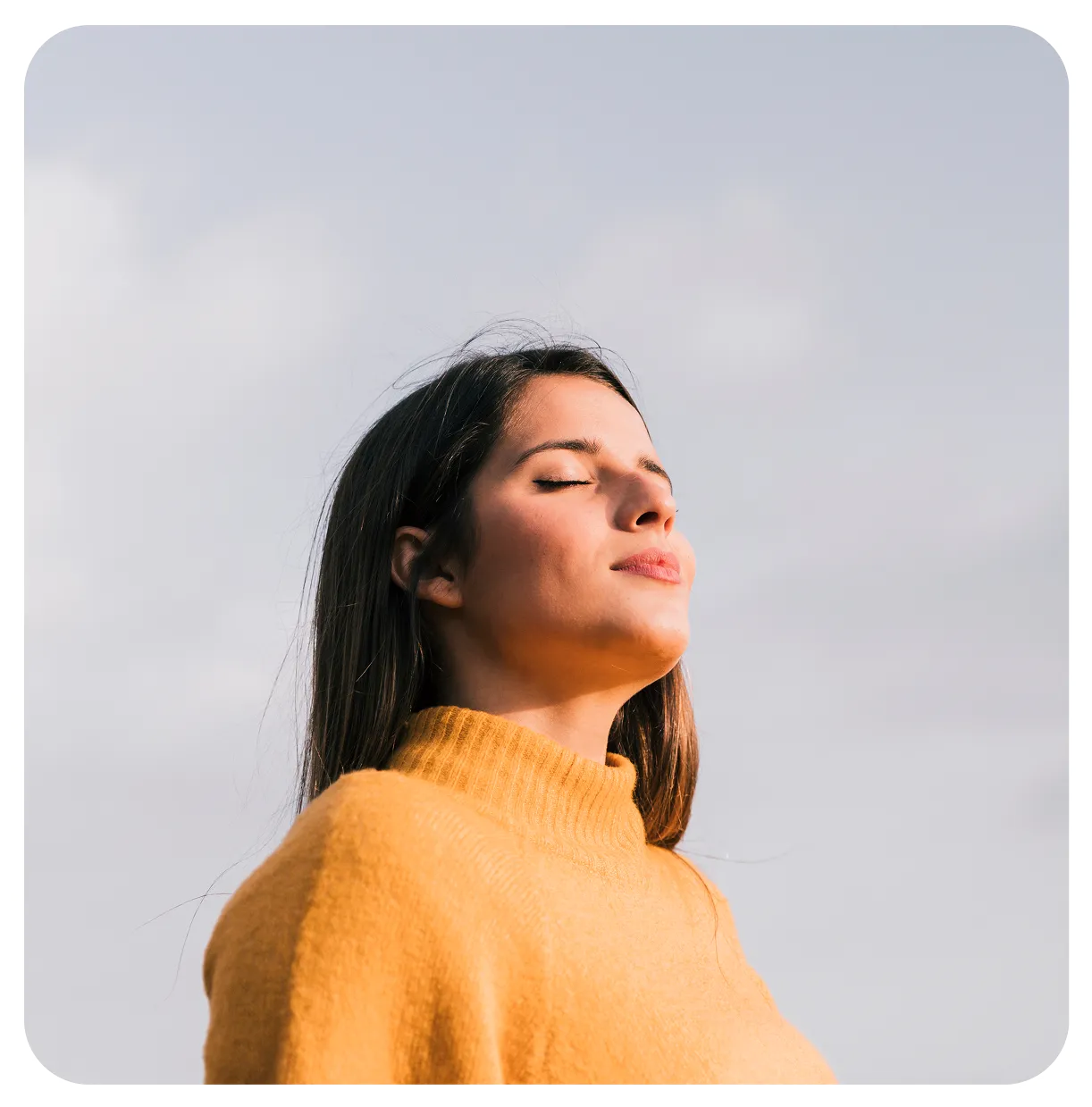 Woman with closed eyes in a mustard yellow sweater enjoying sunlight against a cloudy sky.