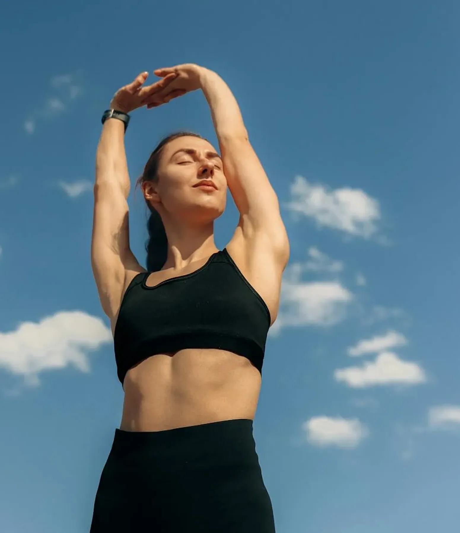 Woman in black workout clothes stretching arms upward with eyes closed against a blue sky with clouds.