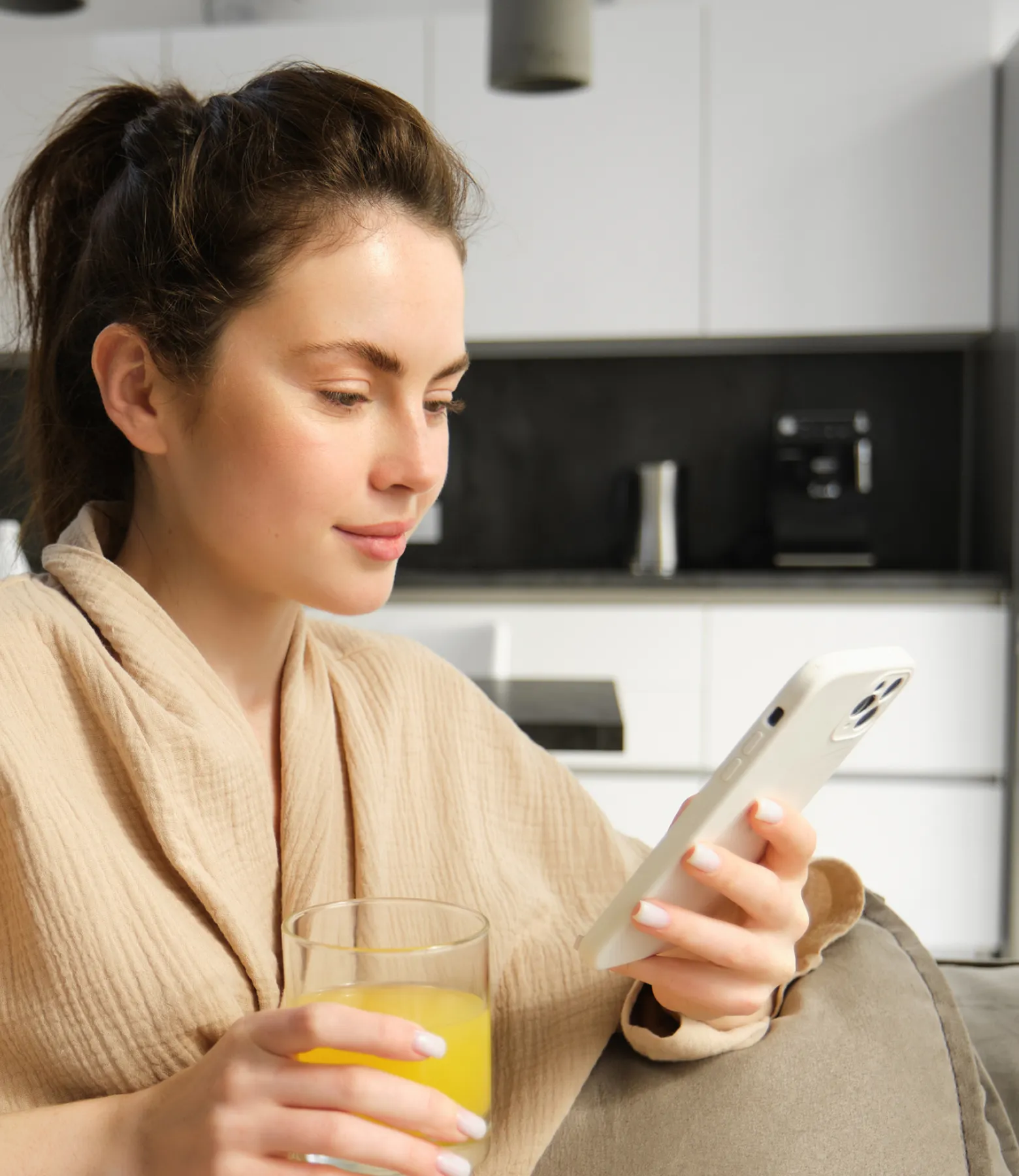 Woman in a beige robe holding a glass of orange juice and looking at her smartphone while sitting on a couch in a modern kitchen.