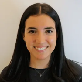 Smiling woman with long dark hair wearing a black top and a small necklace against a plain background.