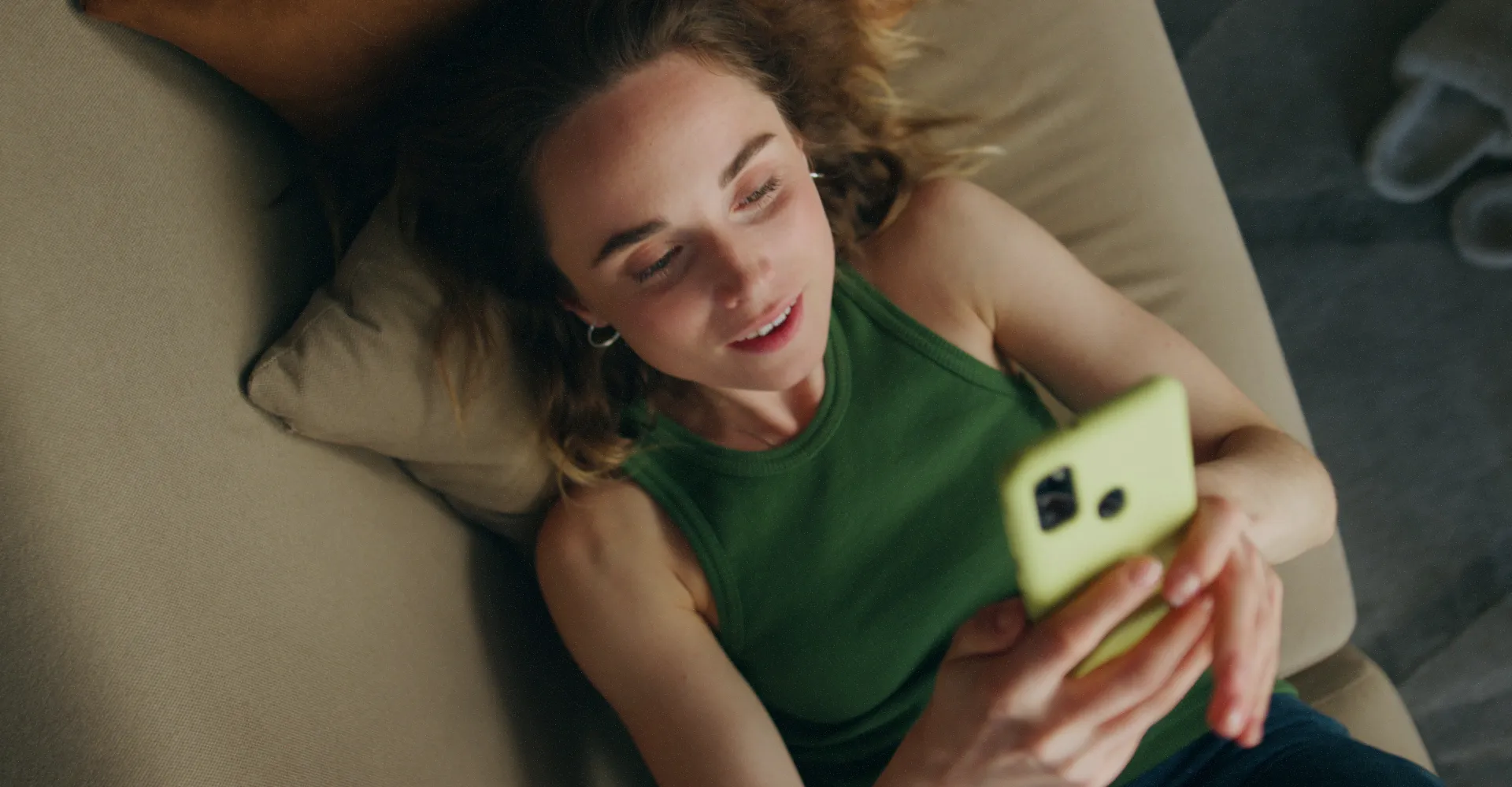 Young woman lying on a beige couch looking at a yellow smartphone with a slight smile.