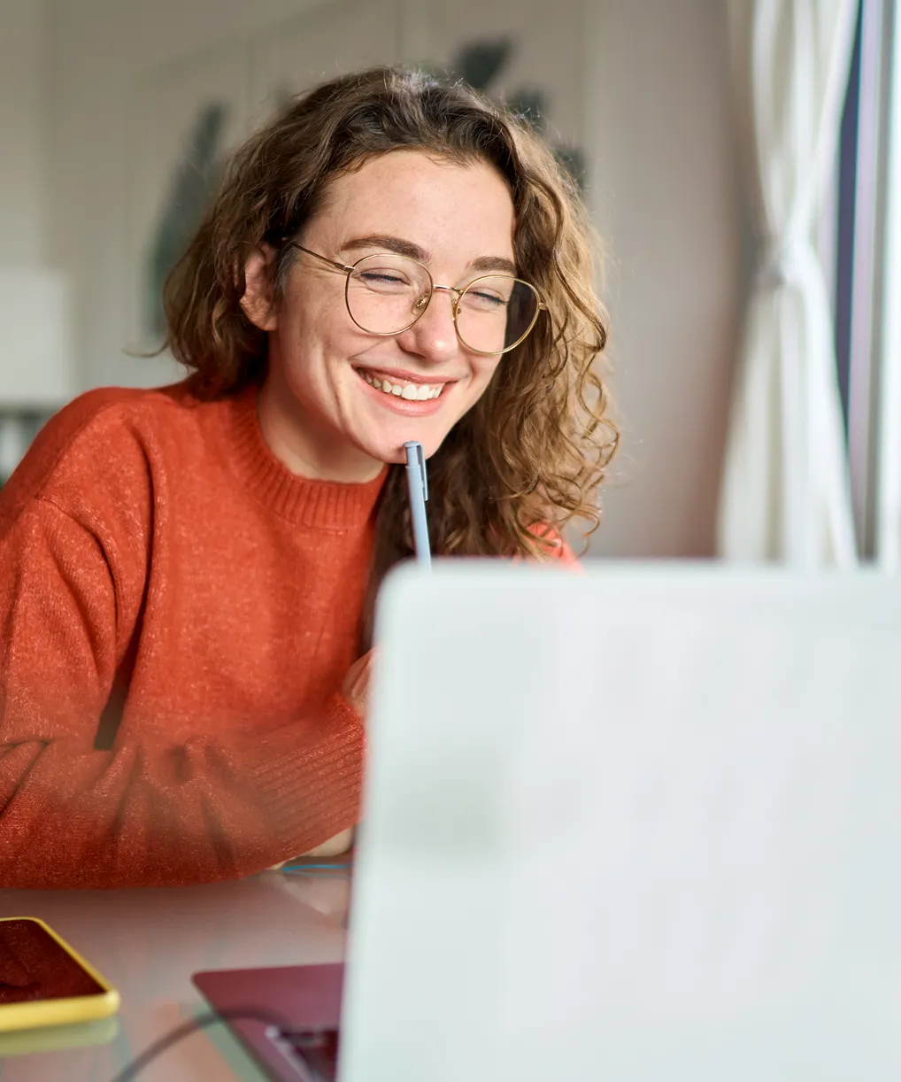 Smiling woman with curly hair and glasses in an orange sweater looking at a laptop screen while holding a pen.