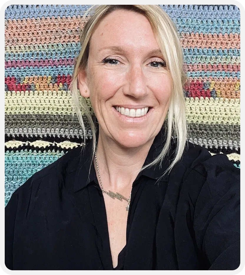 Smiling blonde woman wearing a black shirt and a lightning bolt necklace, sitting in front of a colorful knitted blanket.