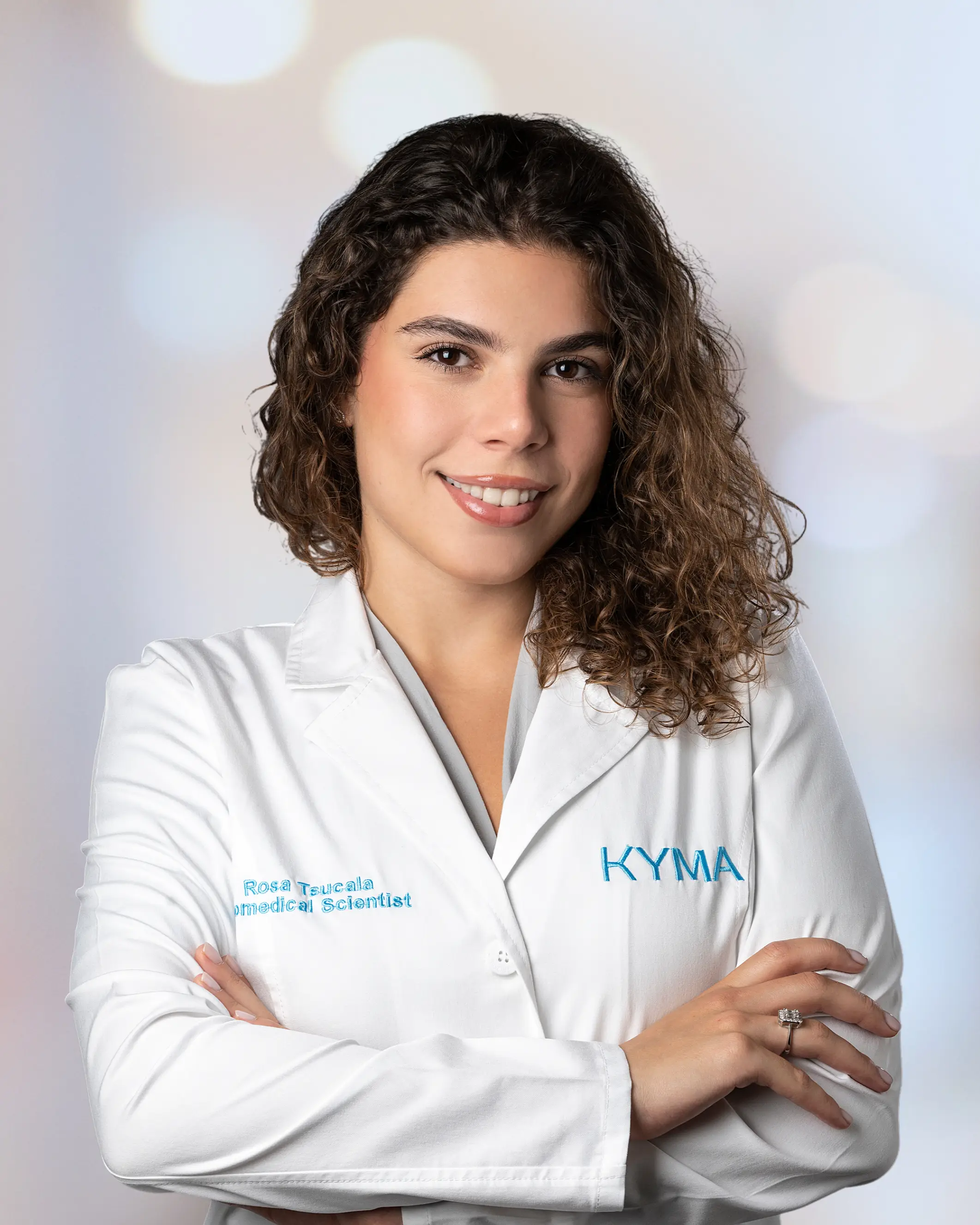 Smiling woman with curly hair wearing a white lab coat labeled KYMA and biomedical scientist.