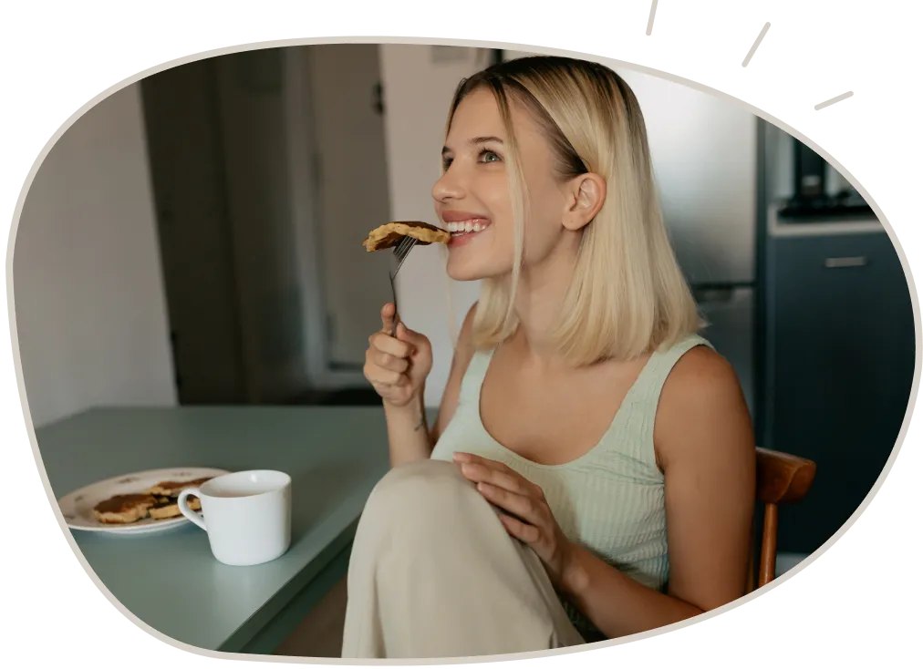 Smiling woman with blonde hair sitting at a table eating a forkful of pancake, with a plate of pancakes and a white mug on the table.
