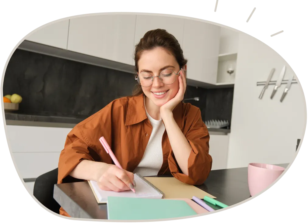 Young woman with glasses sitting at a kitchen table smiling and writing in a notebook with a pink pen.