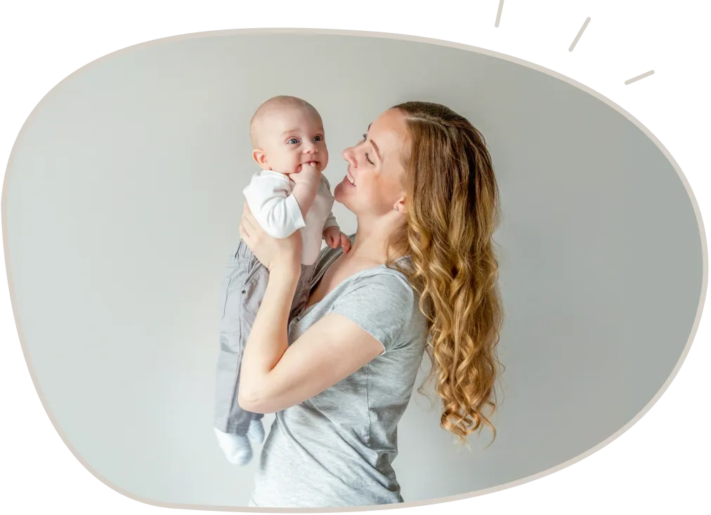 Smiling woman with curly hair holding a baby who is sucking on fingers.