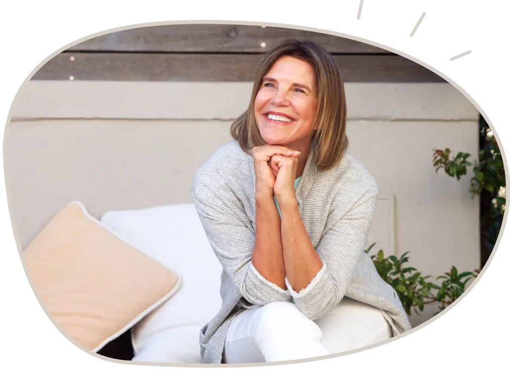 Smiling middle-aged woman sitting outdoors on a cushioned bench with hands clasped under her chin.