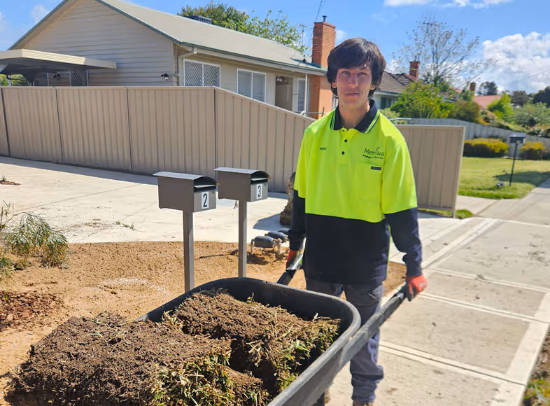A man in fluro standing in front of a house holding a wheel barrow fill of lawn.