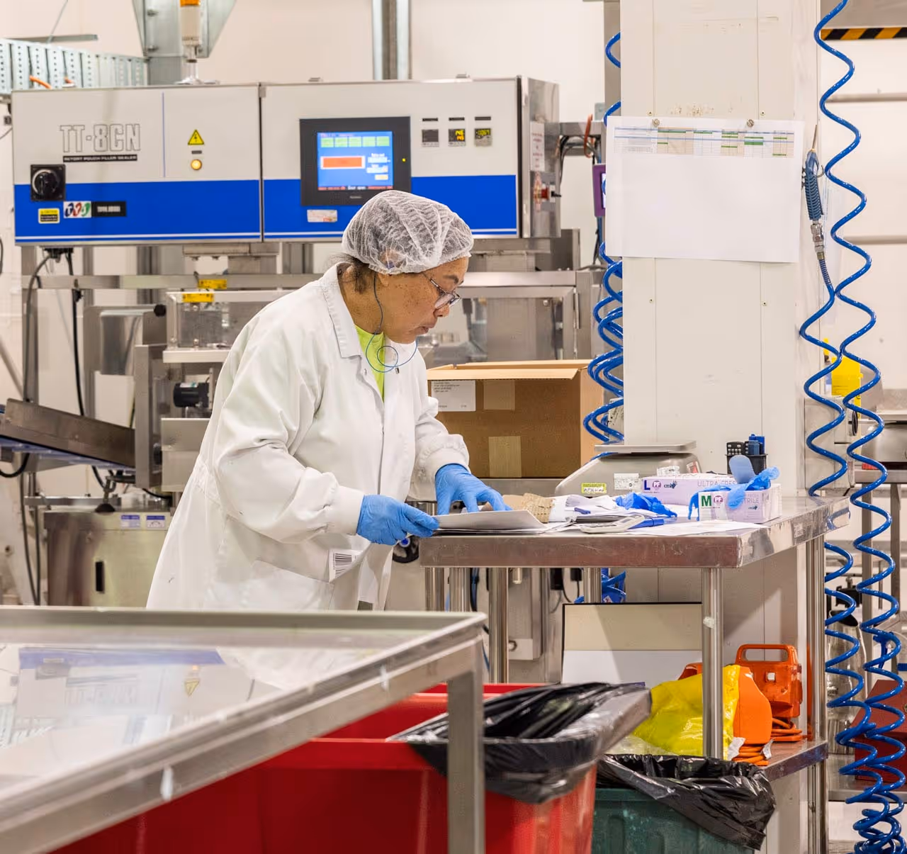 A lady standing in a food room with a coat and hairnet on looking at a table.