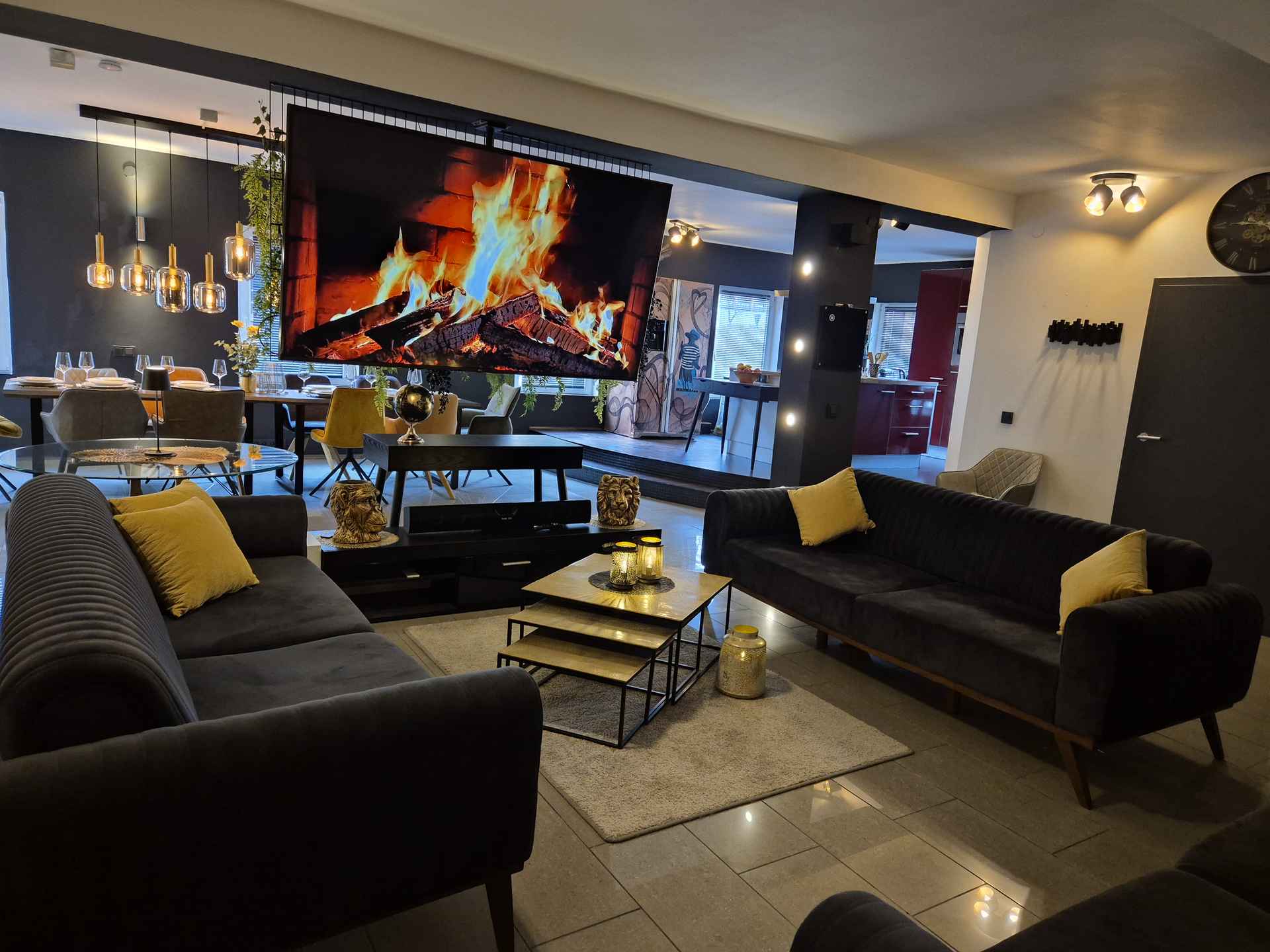 Wohnzimmer mit Kaminfeuer auf einem großen TV, Sofas und ein offenes Wohnen wird gezeigt, Küche und Essbereich im Hintergrund