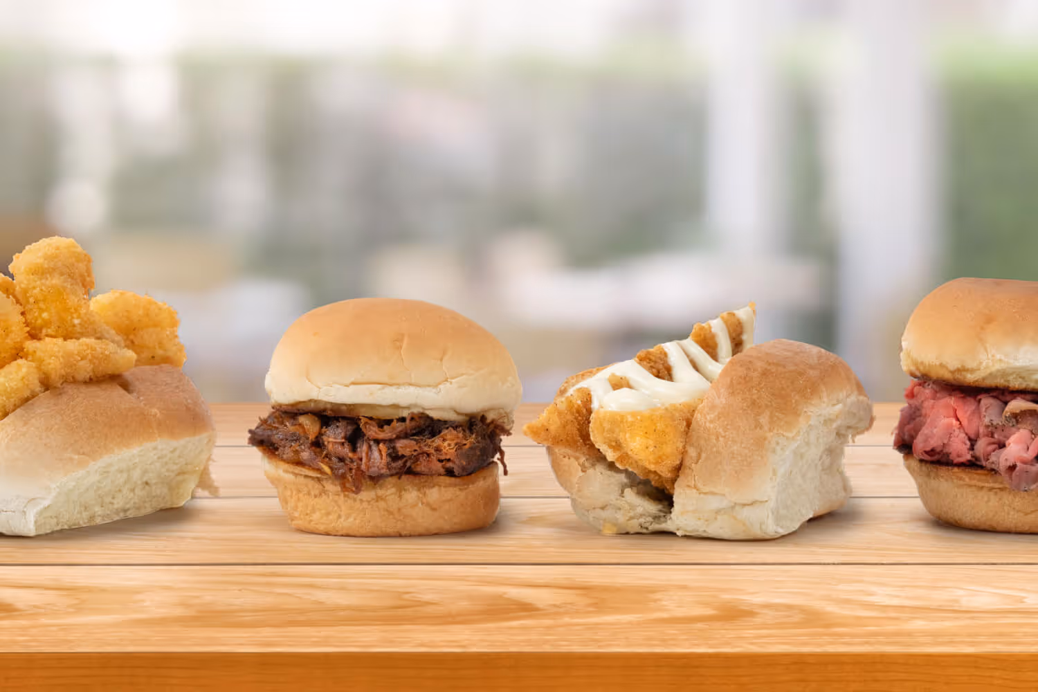 Four sandwiches featuring fried chicken, pulled pork, fish, and roast beef on a wooden surface with a blurred background.