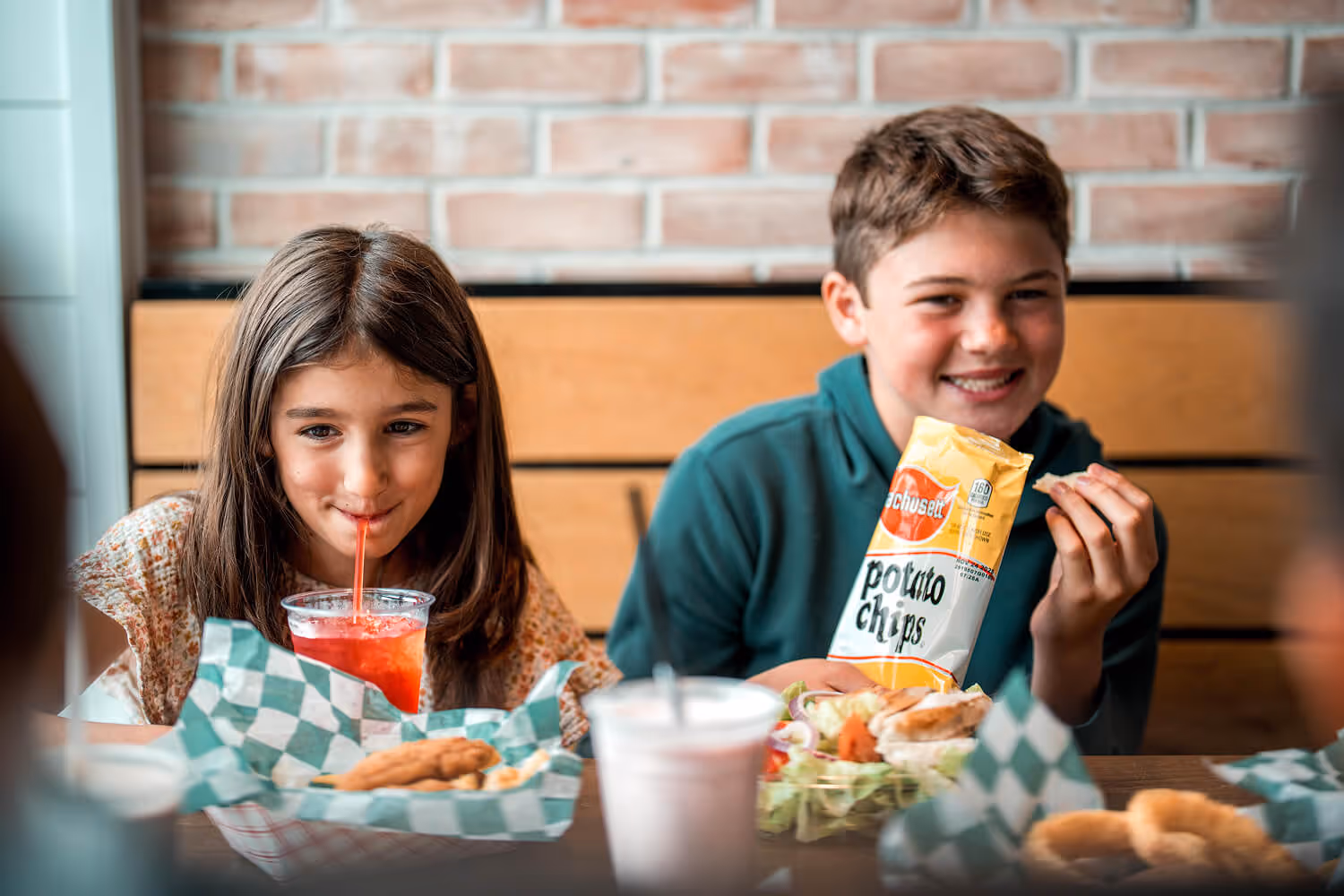 Two children sit at a table with food trays, one drinking a red beverage and the other holding a bag of potato chips.