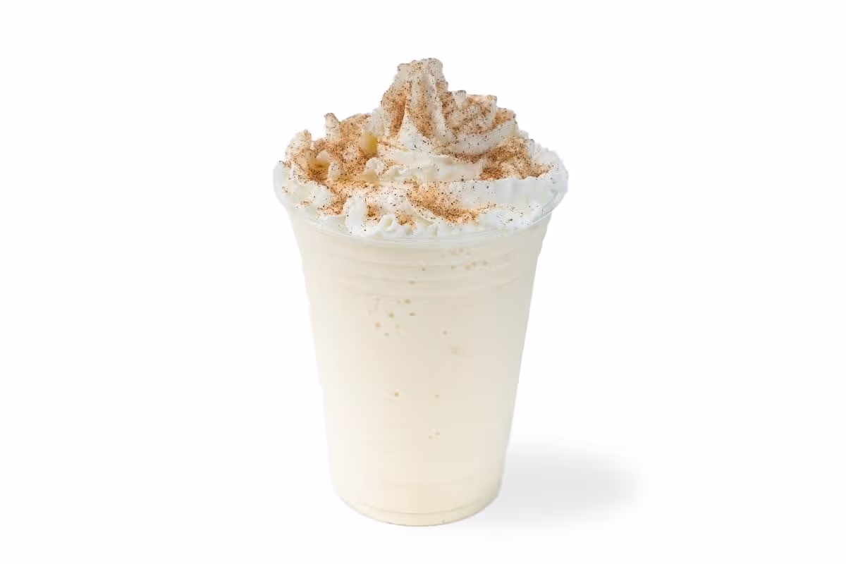 A vanilla milkshake in a clear plastic cup topped with whipped cream and a sprinkle of cinnamon.