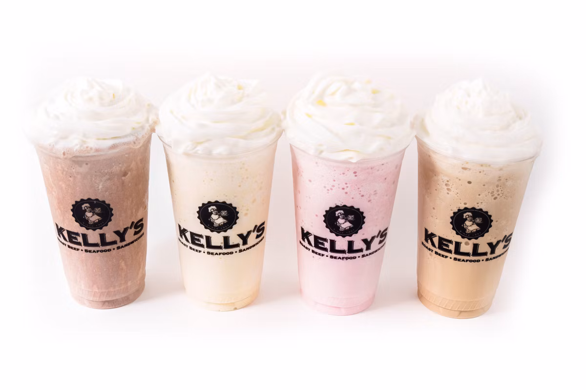 Four milkshakes in clear Kelly’s cups, each topped with whipped cream, lined up on a white background.