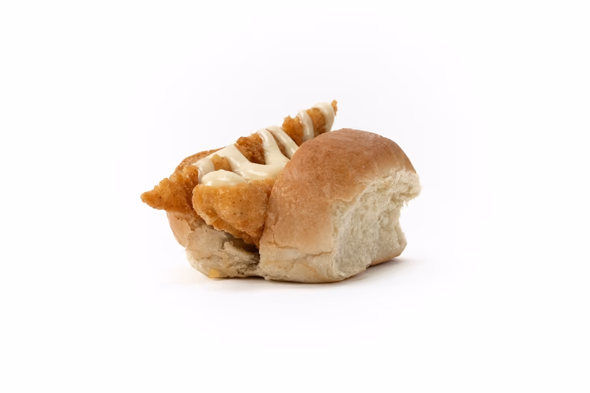 A bread roll with a piece of fried chicken topped with a drizzle of sauce, set against a white background.