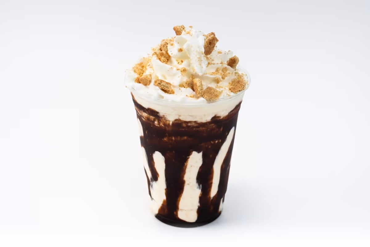 A chocolate milkshake topped with whipped cream and crushed cookie crumbs in a clear plastic cup, isolated on white background.