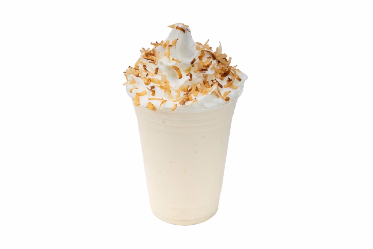 A vanilla milkshake in a clear cup topped with whipped cream and toasted coconut flakes, isolated on a white background.