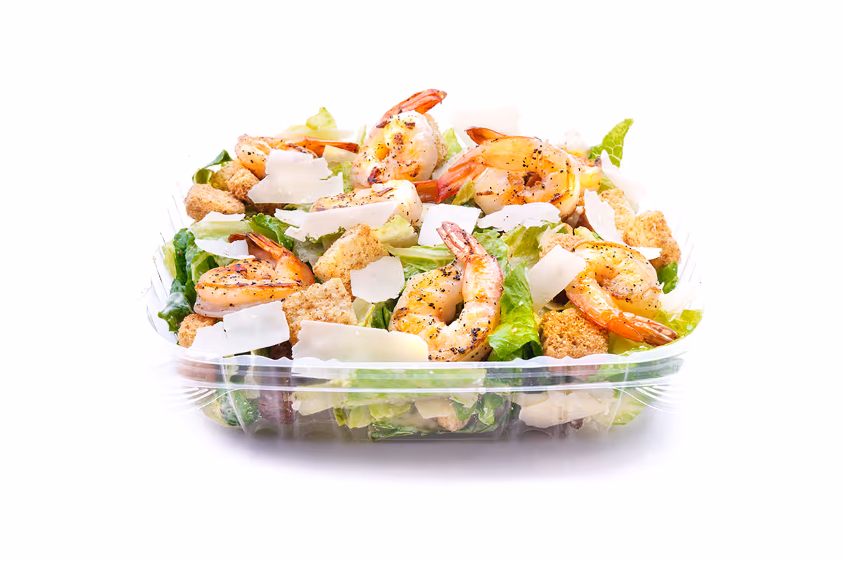 Plastic container of Caesar salad with grilled shrimp, romaine lettuce, croutons, and shaved parmesan cheese on white background.