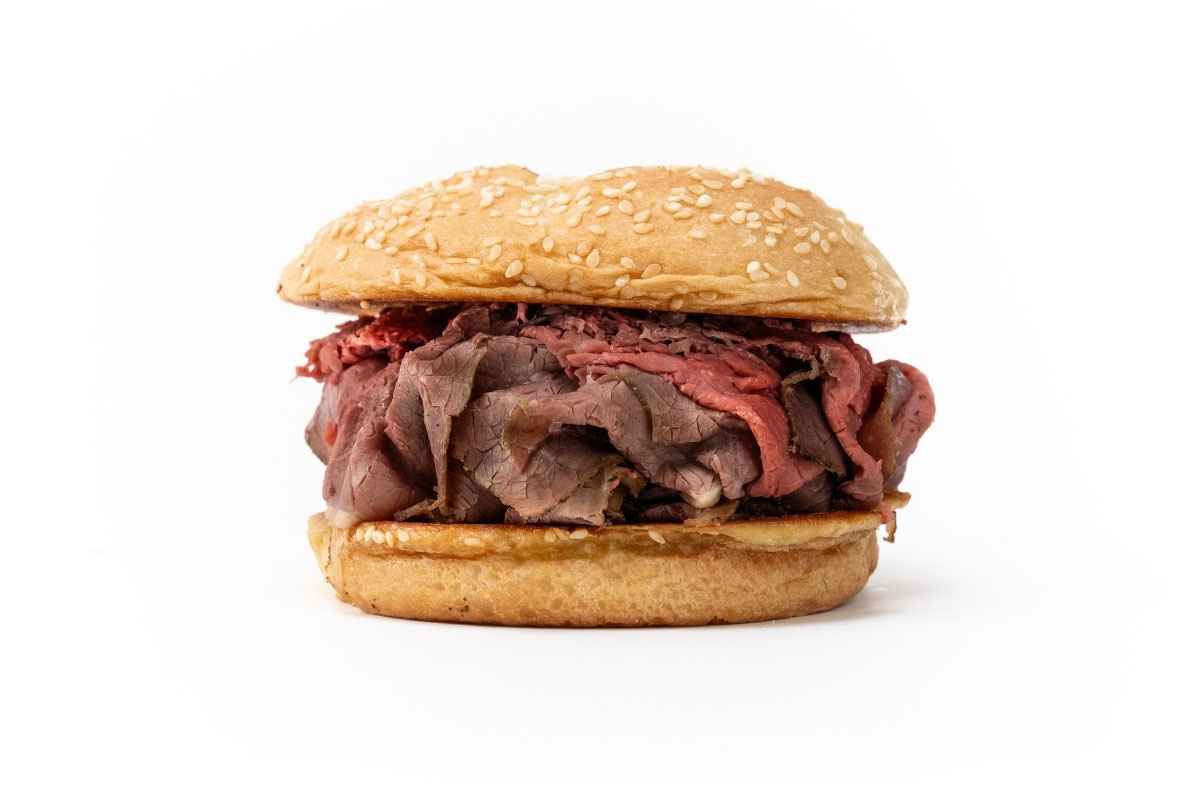 A roast beef sandwich on a sesame seed bun, filled with sliced roast beef, against a white background.
