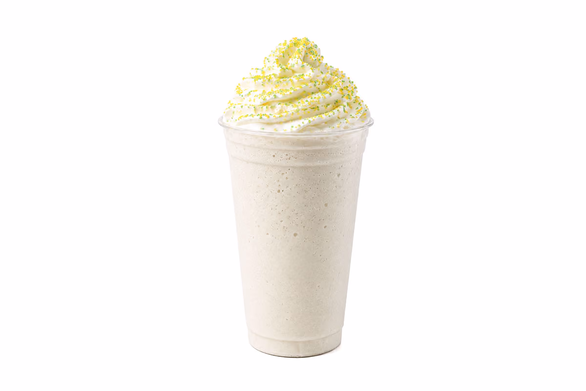 A vanilla milkshake in a clear cup, topped with whipped cream and green sugar sprinkles, on a white background.