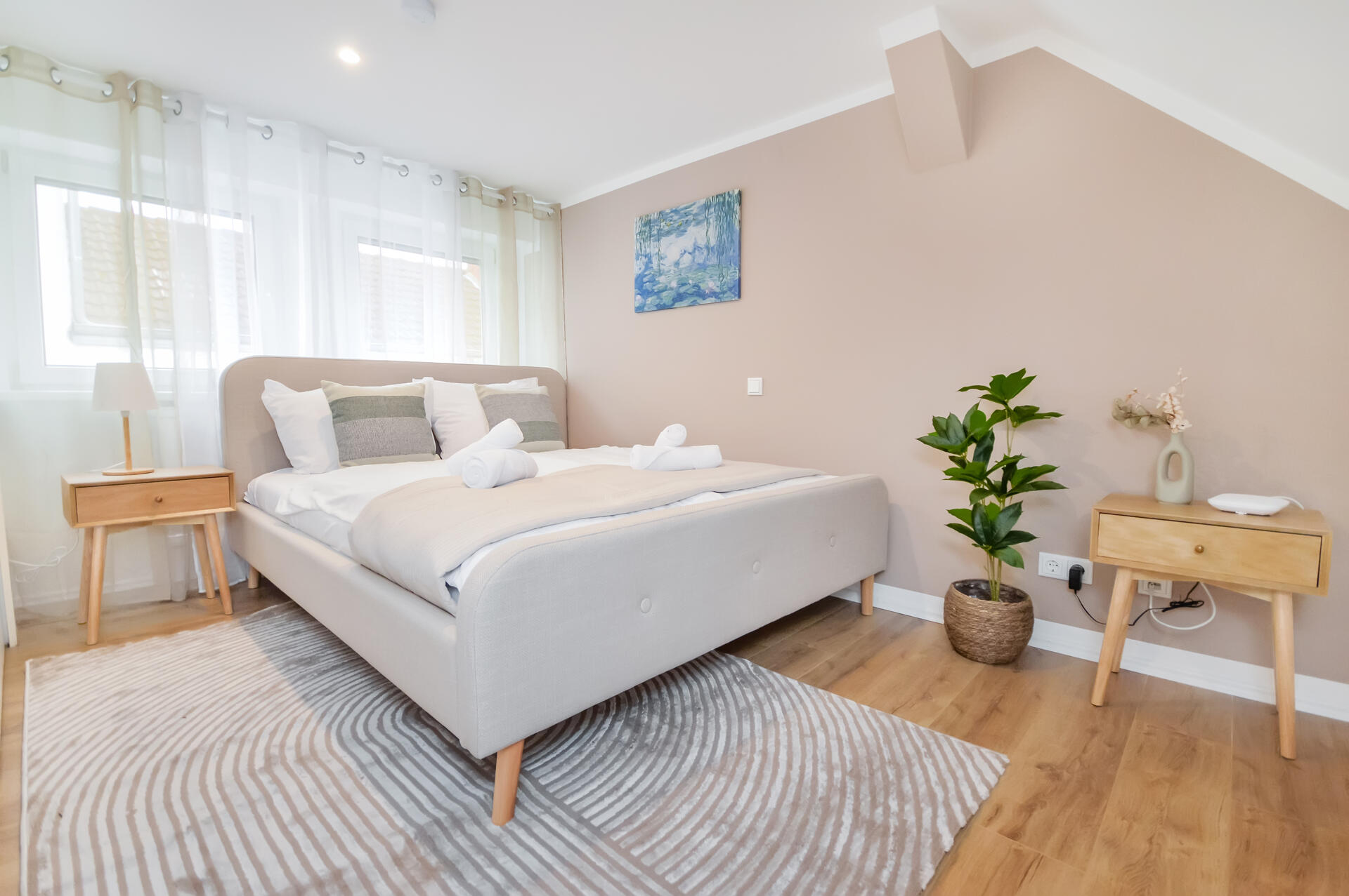 Schlafzimmer mit beigem Akzentwand-Ton, gepolstertem Bett, zwei Holz-Nachttischen mit Lampen, gerollten Handtüchern auf der Decke, großem gemusterten Teppich, Zimmerpflanze im Korb, Laminatboden sowie zwei Fenstern mit hellen Vorhängen.
