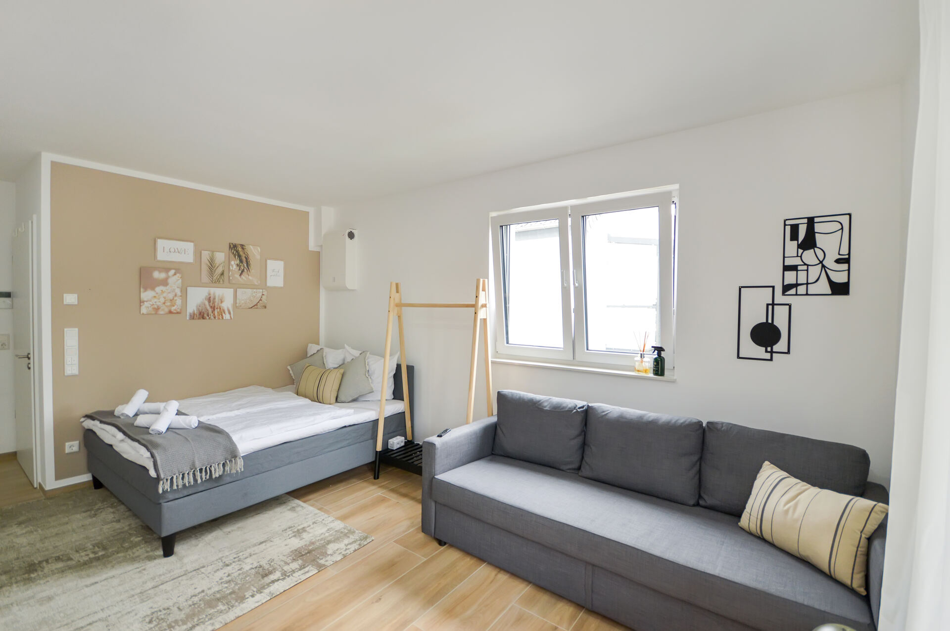 Helles und modern eingerichtetes Studio in den Weinmeister Apartments Bielefeld. Zu sehen sind ein gemütliches Doppelbett mit dekorativen Kissen, ein großes graues Schlafsofa, eine helle beige Akzentwand und moderne Wanddekoration. Der Raum wirkt freundlich und einladend, mit viel Tageslicht durch die großen Fenster – ideal für Kurzzeitvermietung und Geschäftsreisen.