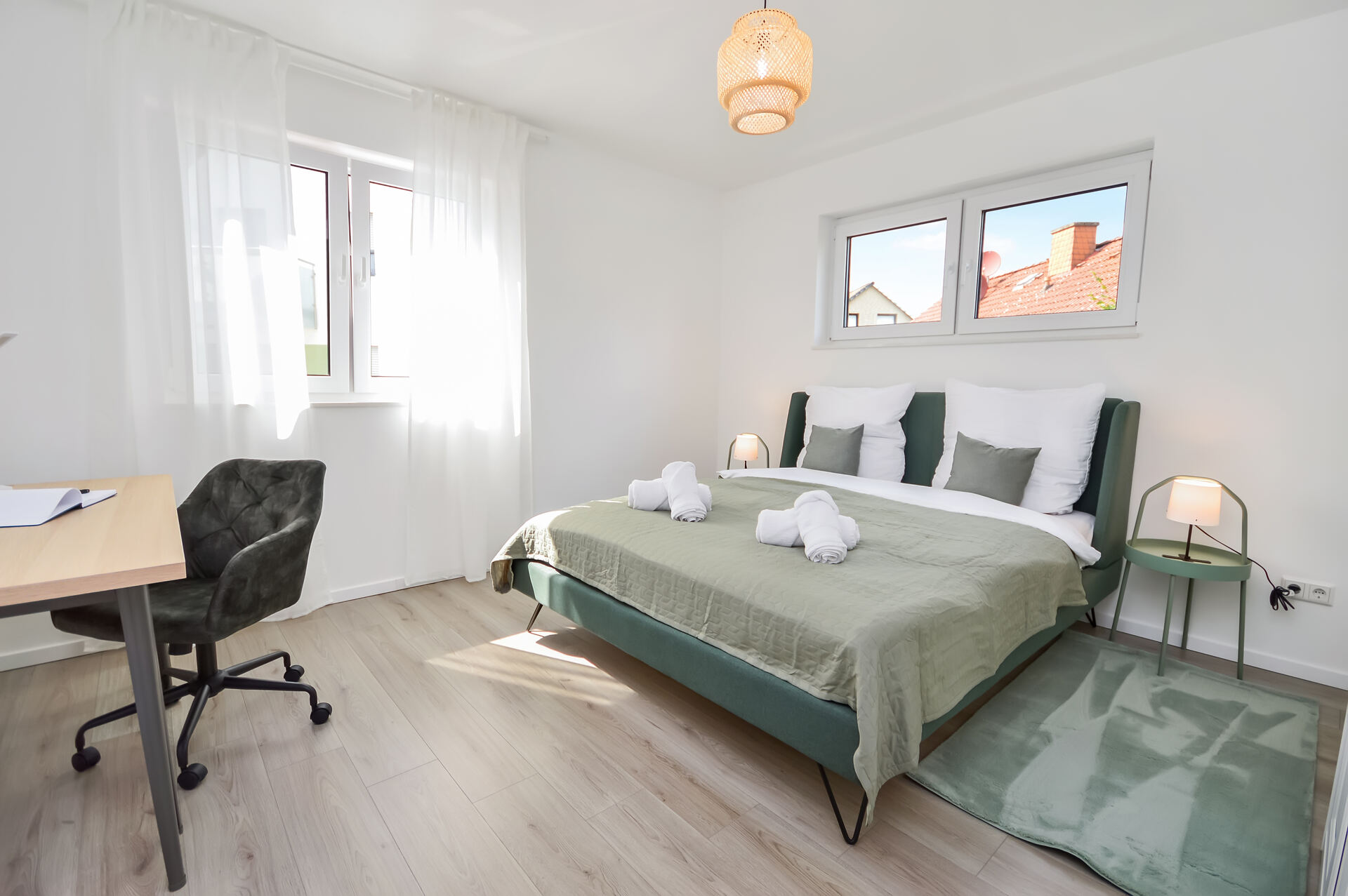 Helles Schlafzimmer in Bielefeld-Brackwede mit großem Doppelbett in Grün, frischer Bettwäsche, modernem Schreibtisch mit Drehstuhl, viel Tageslicht durch zwei Fenster und stilvollen Lampen – ideal für erholsamen Schlaf und komfortables Arbeiten.