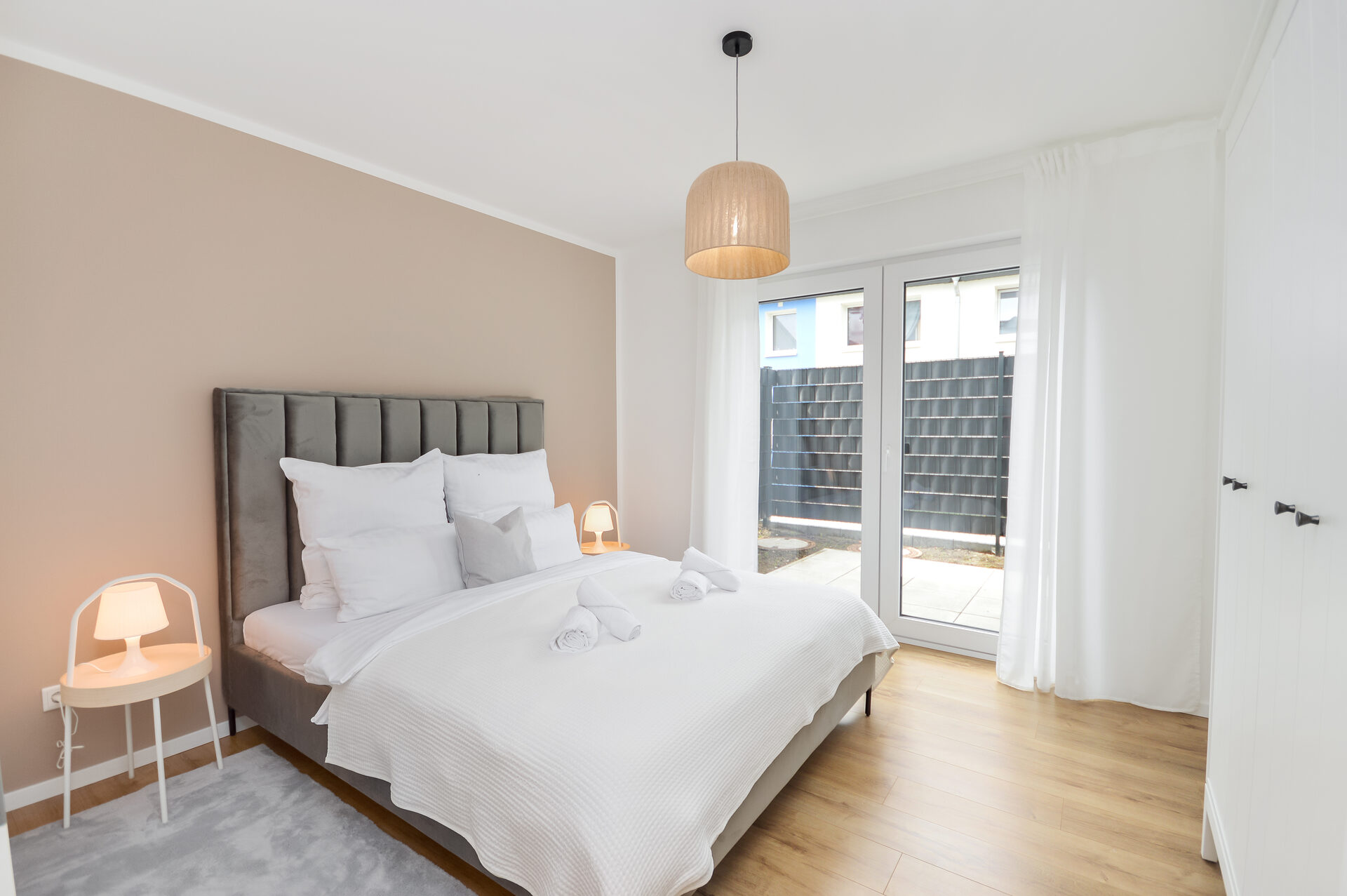 Modernes Schlafzimmer im Erdgeschoss eines Apartments in Bielefeld-Brackwede mit großem Doppelbett, warmen Naturtönen, bodentiefen Fenstern zur Terrasse, weißen Gardinen und hellem Holzfußboden – ideal für erholsame Nächte und viel Tageslicht.