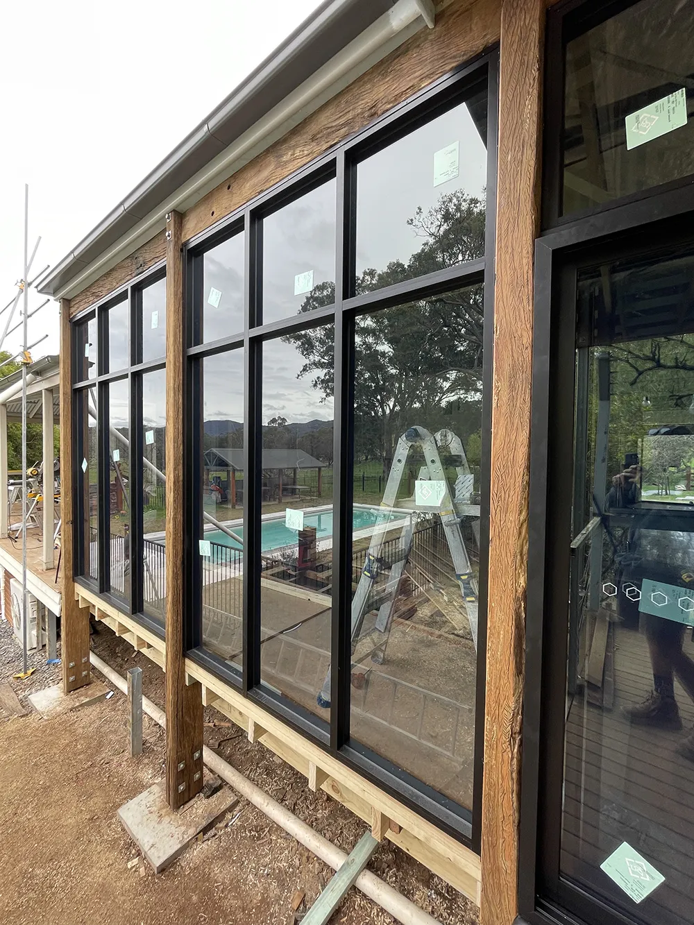 Large black-framed windows installed on a wooden exterior wall of a building under construction, with reflections of trees, a ladder, and a swimming pool.