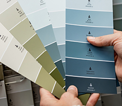 Hands holding paint color swatches ranging from beige to blue shades.