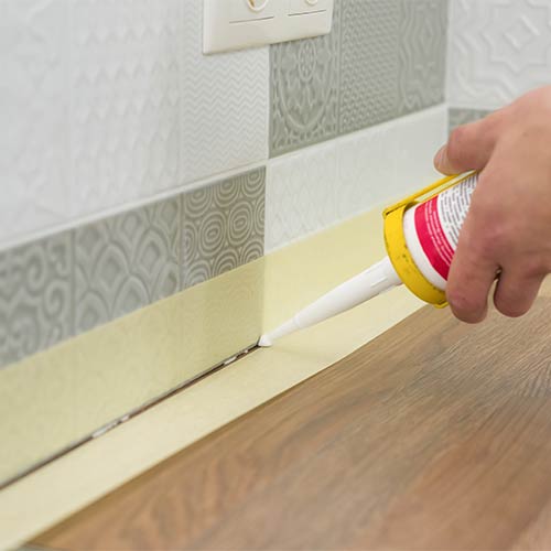 Hand applying white sealant with a caulking gun along the baseboard and wooden floor.