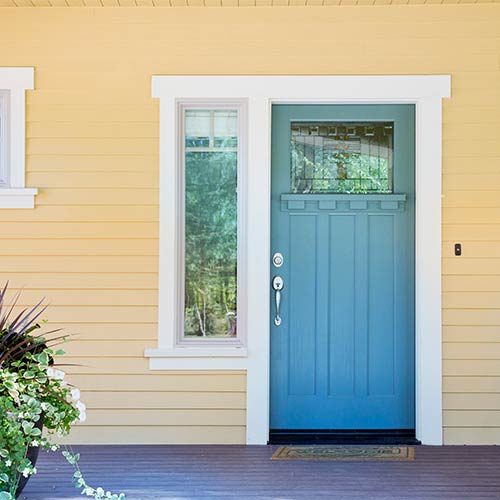 Blue front door with a glass window and decorative trim on a yellow house with vertical siding and a small tall window to the left.
