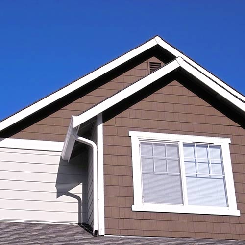 Upper portion of a house showing brown siding with white trim around a window and roof against a clear blue sky.