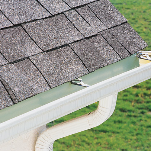 Close-up of a roof with asphalt shingles and a white gutter system with a downspout against a green grassy background.