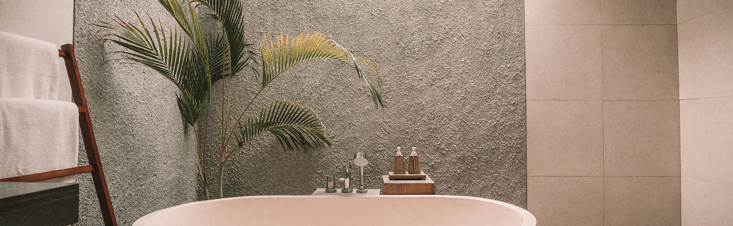 Modern bathroom with a white freestanding bathtub, textured gray wall, palm plant, wooden towel rack with white towels, and soap dispensers on a wooden tray.