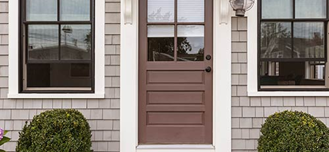 Brown front door with a window panel, flanked by two white-trimmed windows and symmetrical bushes in front.
