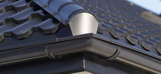 Close-up of a dark-colored roof gutter corner with a downspout attached under a tiled roof.