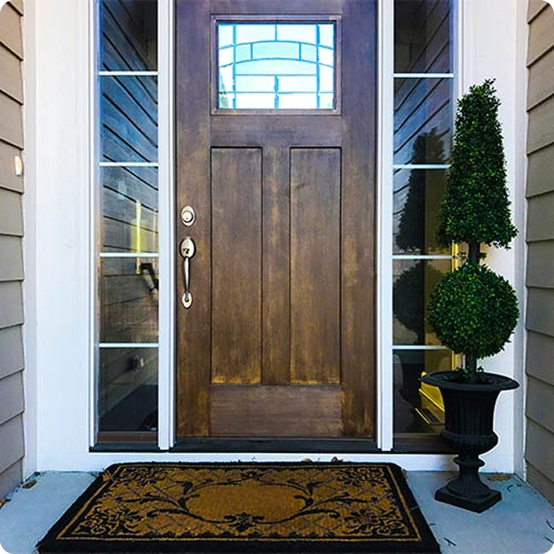 Wooden front door with rectangular window, flanked by glass sidelights, a decorative doormat, and a potted topiary plant.