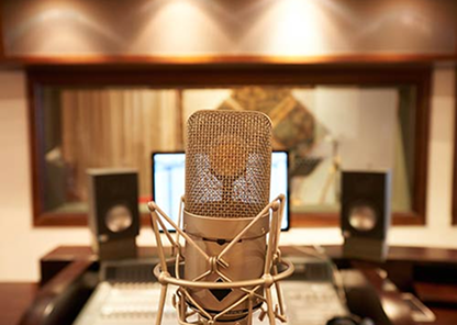 Close-up of a studio microphone with blurred computer monitors and speakers in a recording studio background.