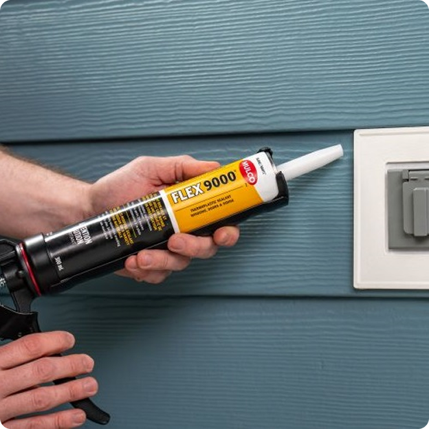 Person holding a tube of FLEX 9000 sealant against a blue textured wall near an electrical outlet.