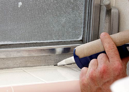 Hand applying white caulk along the edge of a window frame using a caulking gun.