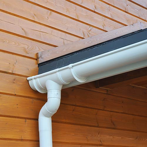 White gutter and downspout installed on a wooden house corner with horizontal planks.