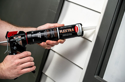Hands applying FLEXTRA sealant with a caulking gun to the exterior siding of a house near a window frame.