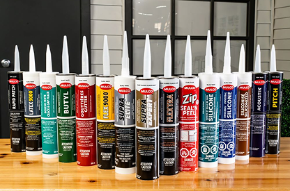 Row of various Mulco branded caulking and adhesive tubes lined up on a wooden surface against a windowed wall.