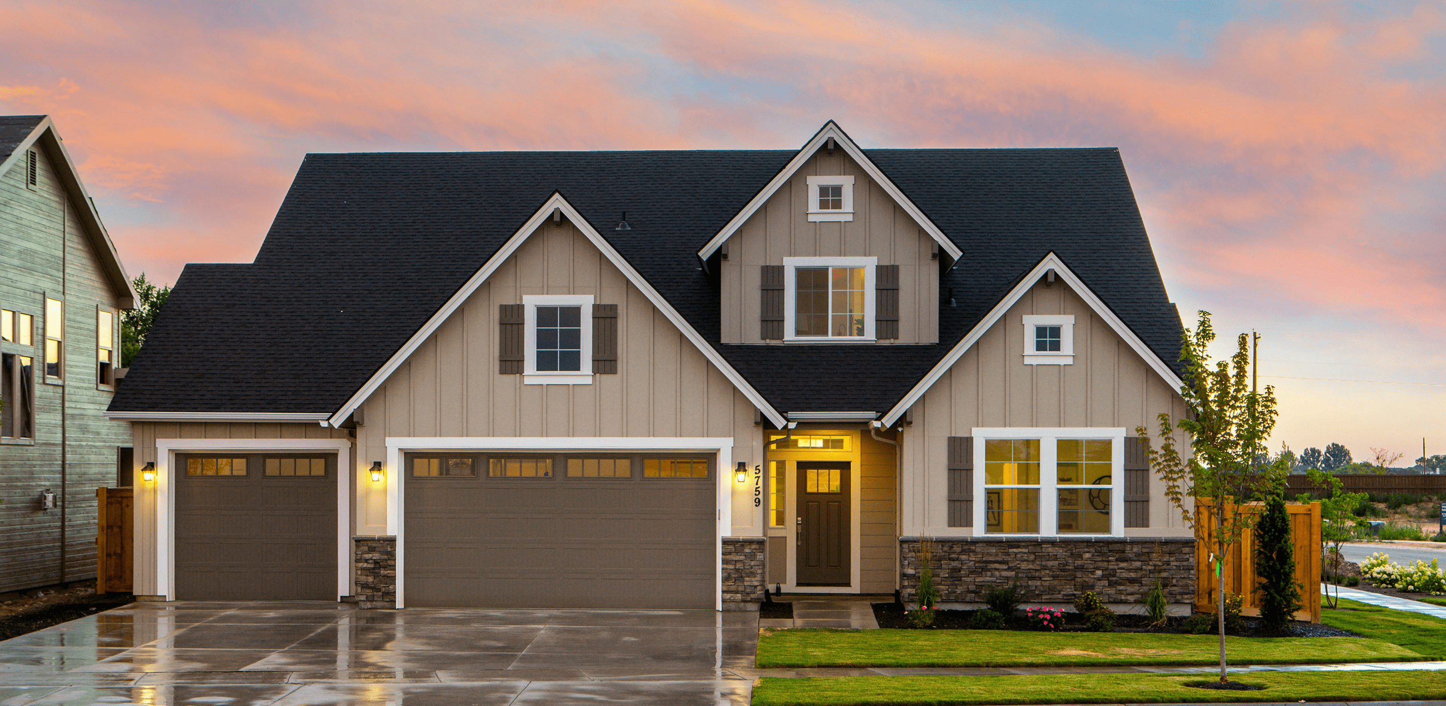 Modern beige house with dark roof, three-car garage, front porch light on, and a manicured lawn at sunset.